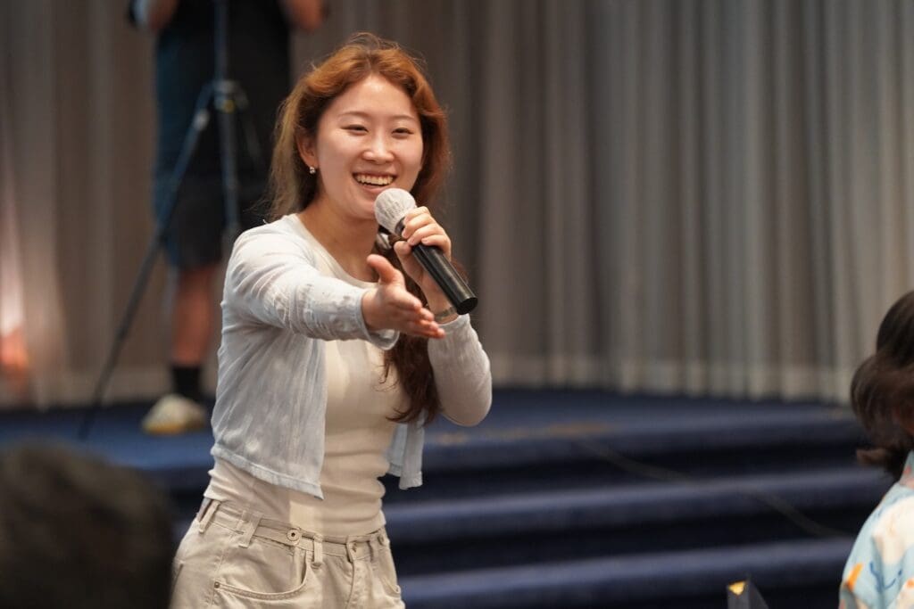 A woman holding a microphone smiles and extends her hand toward the audience, radiating Jeong, while standing in a room with blue carpet and curtains.