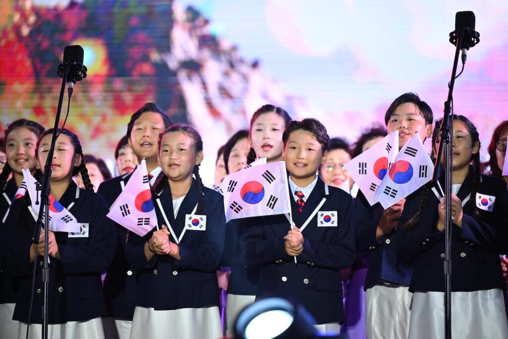 A group of children in uniforms hold South Korean flags and sing on stage at Hangang Festa, with microphones in front of them, a colorful backdrop behind, and the spirit of the Korean Dream shining brightly.
