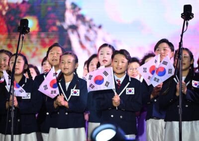 A group of children in uniforms hold South Korean flags and sing on stage at Hangang Festa, with microphones in front of them, a colorful backdrop behind, and the spirit of the Korean Dream shining brightly.