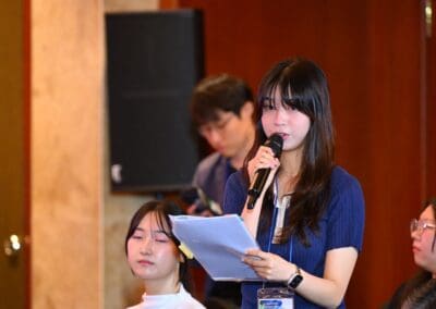 A young woman stands and speaks into a microphone while holding papers at the International Forum on One Korea, with other attendees seated nearby.
