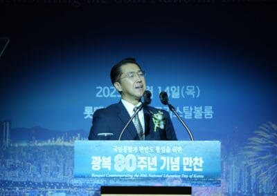 A man in a suit speaks at a podium during a banquet commemorating the 80th National Liberation Day of Korea, with a cityscape and fireworks backdrop, as part of the International Forum on One Korea.