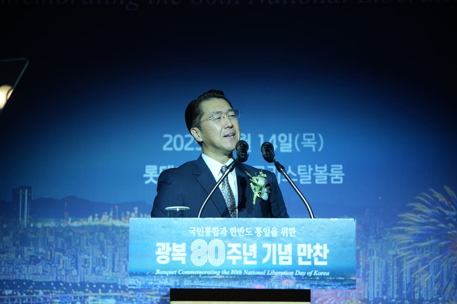A man in a suit speaks at a podium during a banquet commemorating the 80th National Liberation Day of Korea, with a cityscape and fireworks backdrop, as part of the International Forum on One Korea.