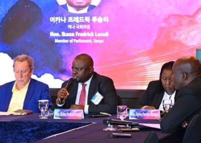 Three people sit at a conference table with microphones and nameplates, speaking in front of a screen displaying "Hon. Ikana Fredrick Lusuli, Member of Parliament, Kenya," at the International Forum on One Korea.