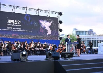 A man and woman speak at a podium onstage during the 2025 Hangang Festa, with an orchestra behind them and a large screen displaying "Opening Declaration," a map of Korea, and the spirit of the Korean Dream.