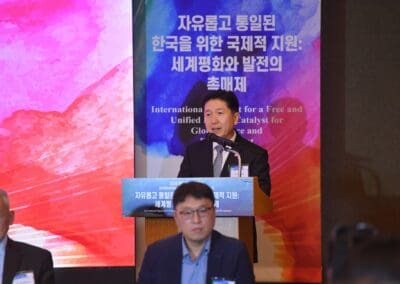 A man in a suit speaks at a podium during the International Forum on One Korea, with a colorful sign in Korean and English displayed behind him.