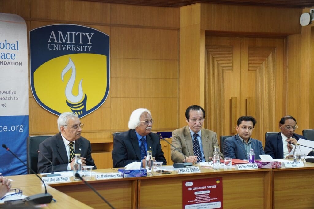 Five men in formal attire are seated at a conference table in front of an Amity University emblem, participating in a panel discussion on Korean Reunification as part of a Global Conversation.