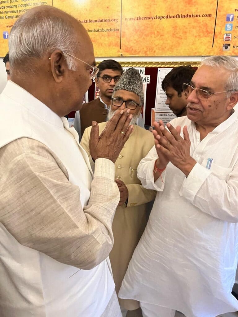 Two elderly men greet each other with folded hands at an event in Rishikesh, while others stand nearby observing. Informational posters about the National Multifaith Coordination Committee are visible in the background.