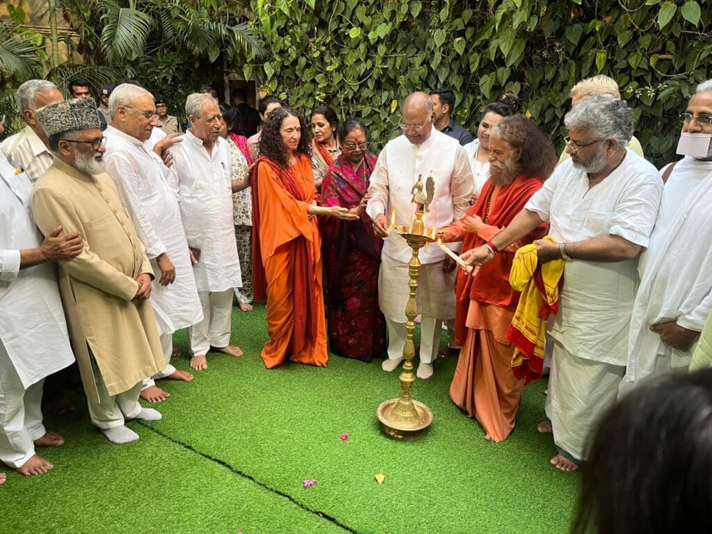 A group of people, dressed mostly in white and traditional Indian attire, gather around a brass oil lamp in an outdoor setting in Rishikesh, participating in a multifaith coordination ceremony for Faith for Development.