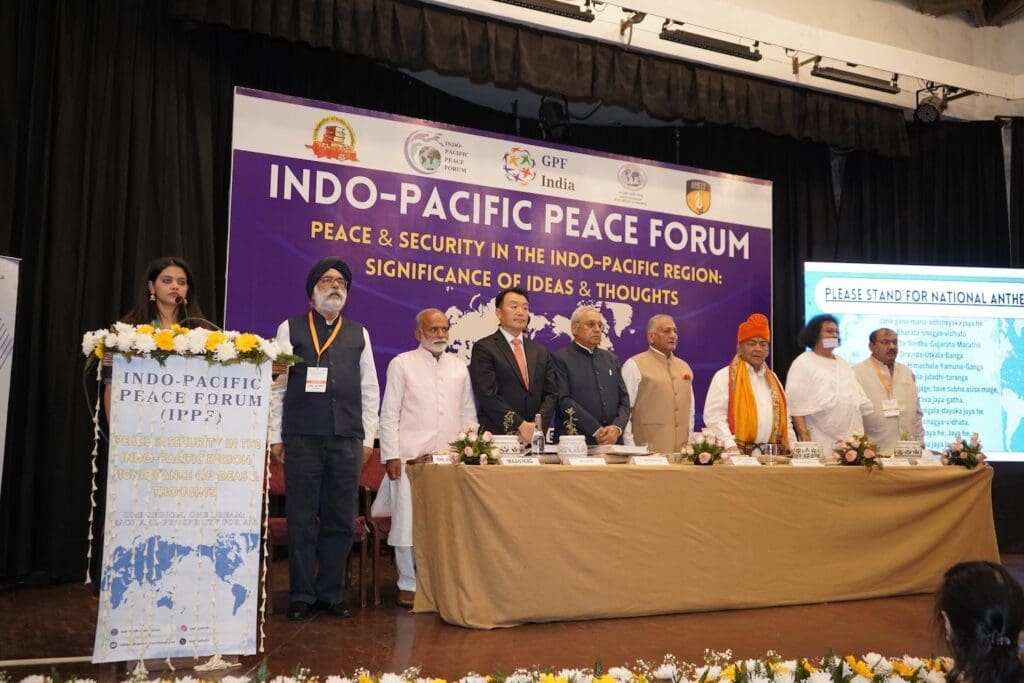 A group of people stands on stage at the Indo-Pacific Peace Forum, with a banner behind them and a woman speaking at a podium about peace and security in the Indo-Pacific region.