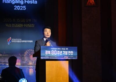 A man in a suit speaks at a podium during an indoor event, with a screen behind him displaying "Hangang Festa 2025" and event details in Korean and English, highlighting the International Forum on One Korea.