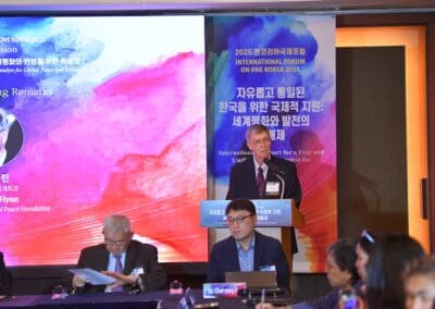 A man stands at a podium delivering a speech at the International Forum on One Korea, with attendees seated and event banners in the background.