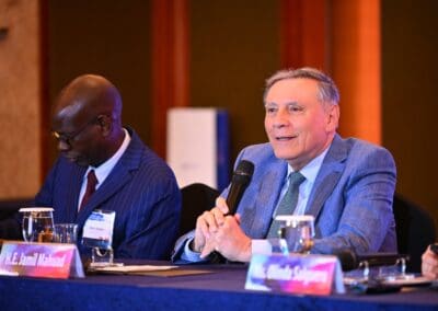 Two men in suits sit at a conference table during the International Forum on One Korea; one speaks into a microphone while the other reads documents. Nameplates and water glasses are visible on the table.