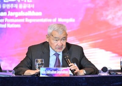 An elderly man in a suit speaks into a microphone at a conference table, with a nameplate in front of him and a colorful background behind, during the International Forum on One Korea.