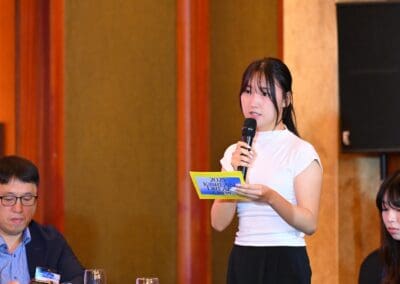 A woman stands holding a microphone and reading from a card while others sit beside her at the International Forum on One Korea, during a formal indoor event.