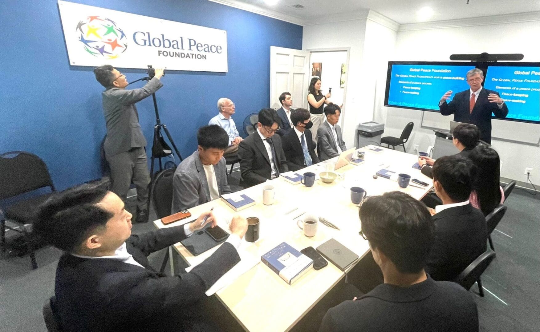 A group of people in business attire sit around a conference table while a presenter speaks near a screen displaying information at the North Korean Young Leaders Assembly, held at the Global Peace Foundation office.