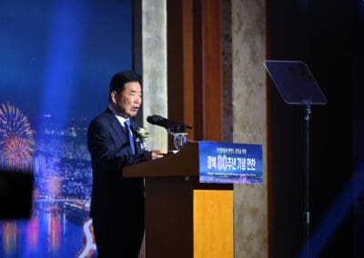 A man in a suit speaks at a podium with microphones during the International Forum on One Korea, with a cityscape and fireworks backdrop behind him.