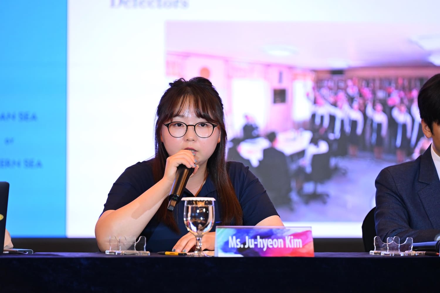 At an International Forum on One Korea in 2025, a woman with glasses speaks into a microphone at a panel, seated behind a nameplate reading
