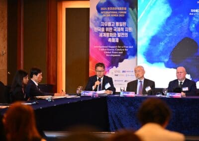 Four men in suits sit at a panel table with microphones during the One Korea 2025 International Forum. A blue banner with bilingual text is displayed in the background.