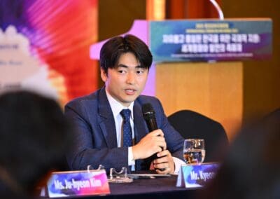 At the International Forum, a man in a suit speaks into a microphone at a conference table on Day 2, with a nameplate reading "Mr. Kyeong" in front of him and a colorful One Korea 2025 banner in the background.
