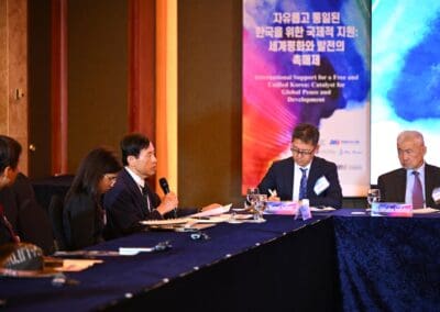 Five people in formal attire sit at a conference table, engaged in discussion. A banner reading "One Korea 2025 International Forum" is visible in the background, indicating the event is on Day 2.