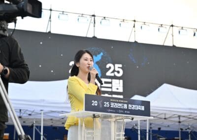 A woman in a yellow jacket speaks into a microphone at an outdoor event, standing behind a podium with a sign reading "2025 Korea Ondream Community Festival," celebrating the spirit of the Korean Dream.