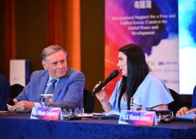A man in a blue suit and a woman in a white blouse speak into microphones at a conference table with nameplates and an International Forum on One Korea event banner in the background.