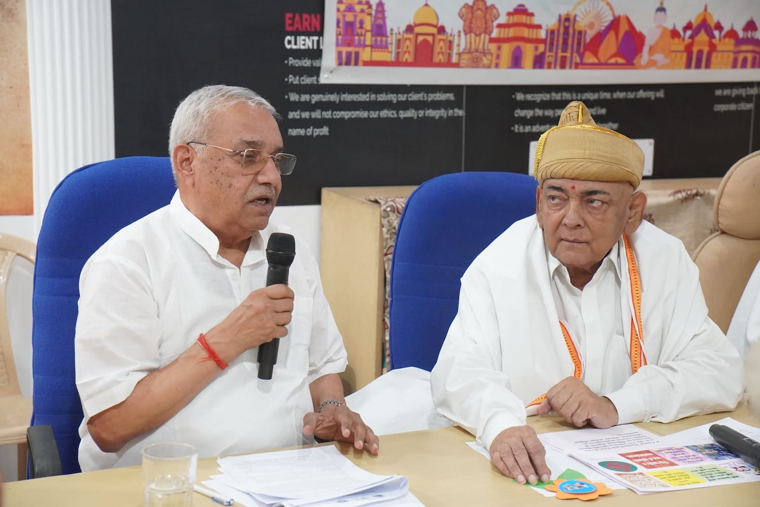 Two elderly men sit at a table; one speaks into a microphone while the other, wearing a traditional cap, holds papers. A colorful poster marking the International Day of Families in India is visible in the background.