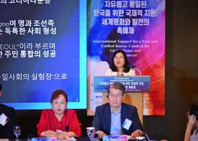 A woman speaks at a podium during the International Forum on One Korea, with two seated panelists in front and banners featuring Korean and English text displayed in the background.