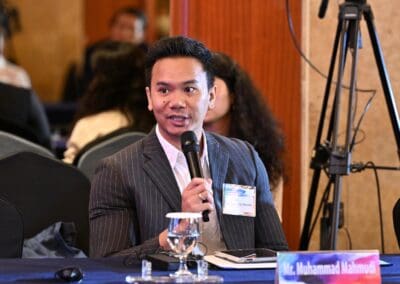 A man in a suit speaks into a microphone at a conference table during the International Forum, with a nameplate labeled "Mr. Muhammad Mahmudi" in front of him, highlighting the One Korea initiative for 2025.