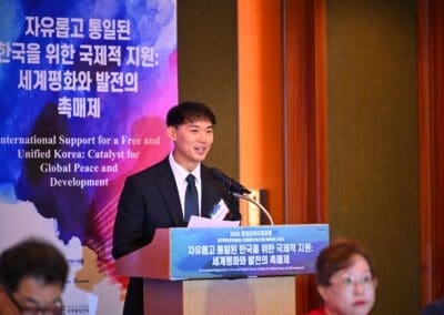 A man in a suit speaks at a podium during the International Forum on One Korea, with a bilingual banner and a woman seated nearby.