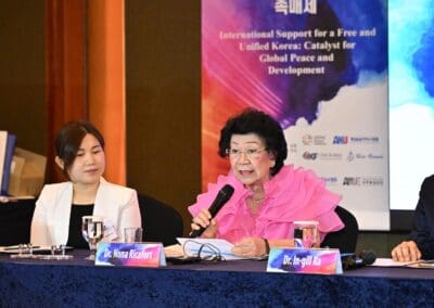 Two women sit at a conference table; one, speaking into a microphone and wearing a bright pink ruffled blouse. Behind them, a banner for the 2025 International Forum on global peace and One Korea unification is visible.