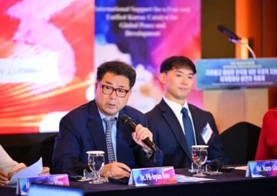 Two men in suits sit at a conference table with microphones and water glasses; one man speaks while the other listens. A sign for the International Forum on One Korea and a colorful backdrop are visible behind them.