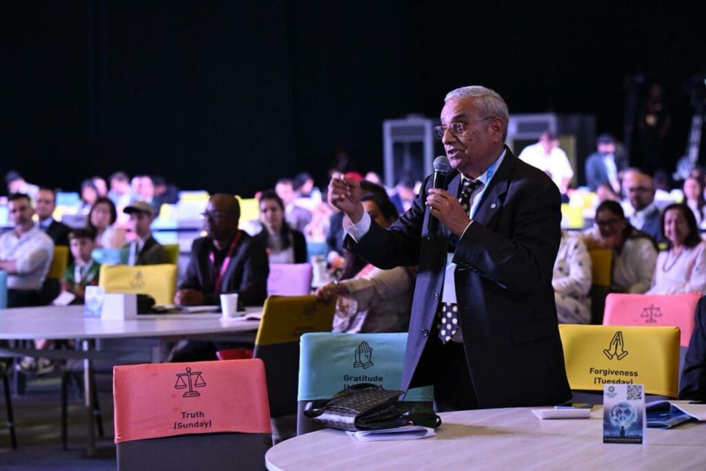 At the Peace Summit 2025, a man in a suit speaks into a microphone while standing among seated attendees at a conference with colored chair covers labeled with virtues.