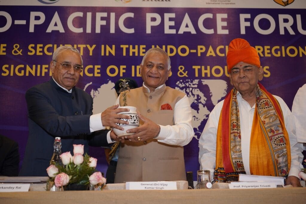 Three men in formal attire stand at a table; the man in the center receives a potted plant from the man on the left. A banner about the Indo-Pacific Peace and Security Forum is visible behind them, highlighting hopes for a peaceful future.