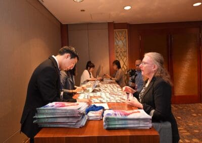 A woman speaks with a man at an International Forum on One Korea registration desk covered with magazines and conference badges; several people work at laptops in the background.