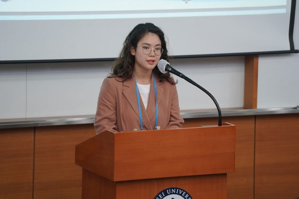 A woman wearing glasses and a brown blazer stands at a podium, speaking into a microphone about Korea reunification in a classroom or conference setting.