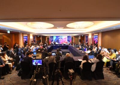 A formal conference at the International Forum on One Korea with many attendees seated around a large U-shaped table, cameras recording, and a presentation screen at the front of the room.