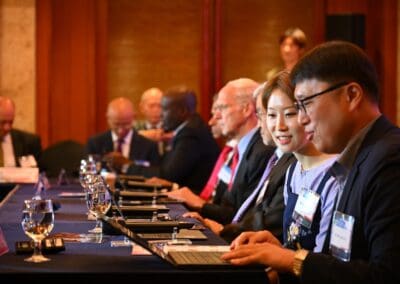 A group of people in business attire sit at a conference table using laptops, with glasses of water and name tags visible at the International Forum on One Korea.