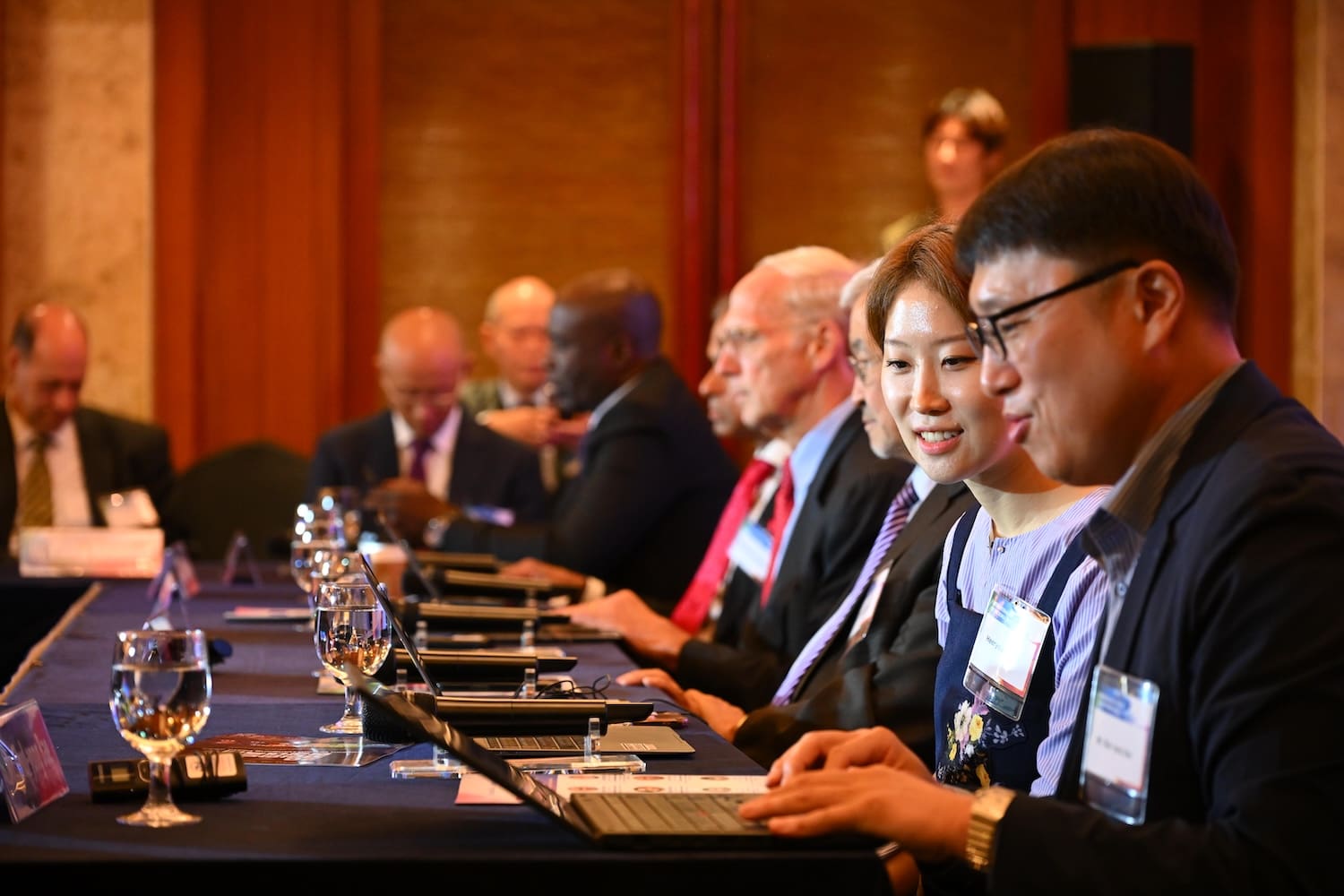 A group of people in business attire sit at a conference table using laptops, with glasses of water and name tags visible at the International Forum on One Korea.