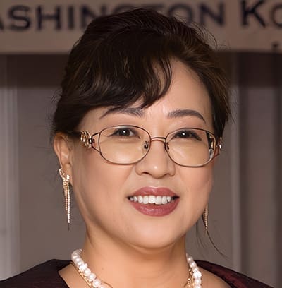 S3 Role of Korean Diaspora 3. Yu-sook Kim Dr. Pil-hyun Koo, a woman with glasses, pearl necklace, and earrings, smiles at the camera; she has dark hair styled up and is wearing formal attire.