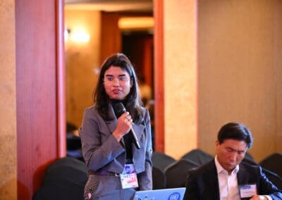 A woman with a staff badge speaks into a microphone while standing at the International Forum on One Korea; a man sits nearby looking down.