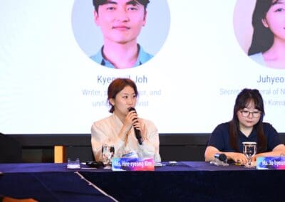 Two women sit at a table with microphones and nameplates, one speaking into a microphone, during an International Forum panel discussion. Large photos and names are displayed on a screen behind them at One Korea 2025.