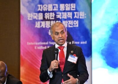 A man in a suit speaks into a microphone at an event. A colorful banner with Korean and English text, featuring the International Forum on One Korea, is displayed in the background.