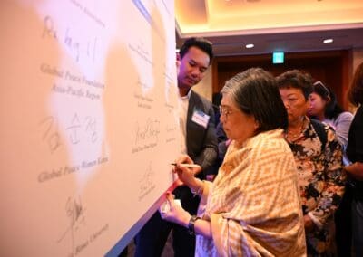 At an indoor event, a woman signs a large white board alongside others at the 2025 International Forum for One Korea, with several signatures already visible on the board.