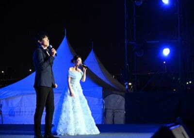 A man in a suit and a woman in a blue gown are singing on an outdoor stage at night, illuminated by blue lights, celebrating the spirit of Korean Dream at Hangang Festa 2025.