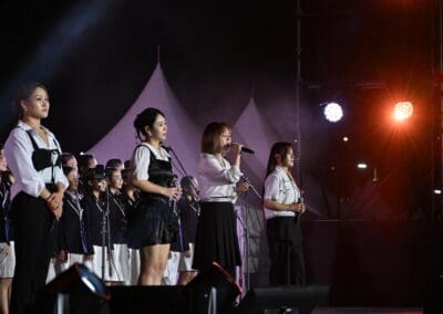 A group of women sings on stage in coordinated black and white outfits under bright stage lights at Hangang Festa 2025, with a choir and tents visible in the background, capturing the spirit of the Korean Dream.