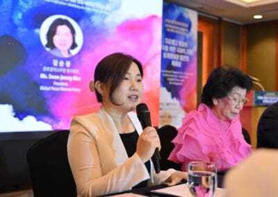 A woman speaks into a microphone at a conference table during the 2025 One Korea International Forum, with another woman seated beside her and a presentation screen in the background.