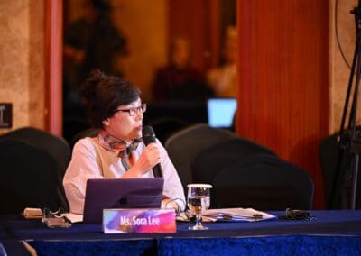 Ms. Sora Lee speaks into a microphone while seated at a table with a laptop, documents, and a glass of water during the International Forum on One Korea.
