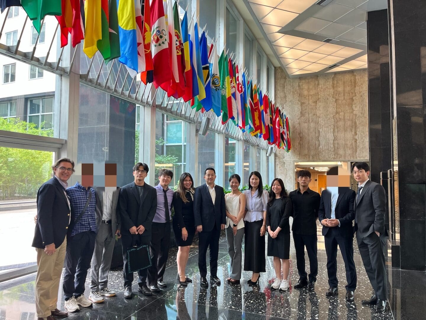 A group of people in business attire stand together indoors beneath a row of international flags and large windows, gathered for the North Korean Young Leaders Assembly.
