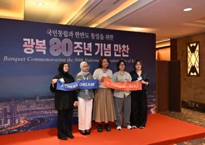Five people stand indoors holding ribbons that say "Korean Dream" and "One Dream One Korea One World" in front of a backdrop for the 80th National Liberation Day of Korea banquet, part of the International Forum on One Korea.